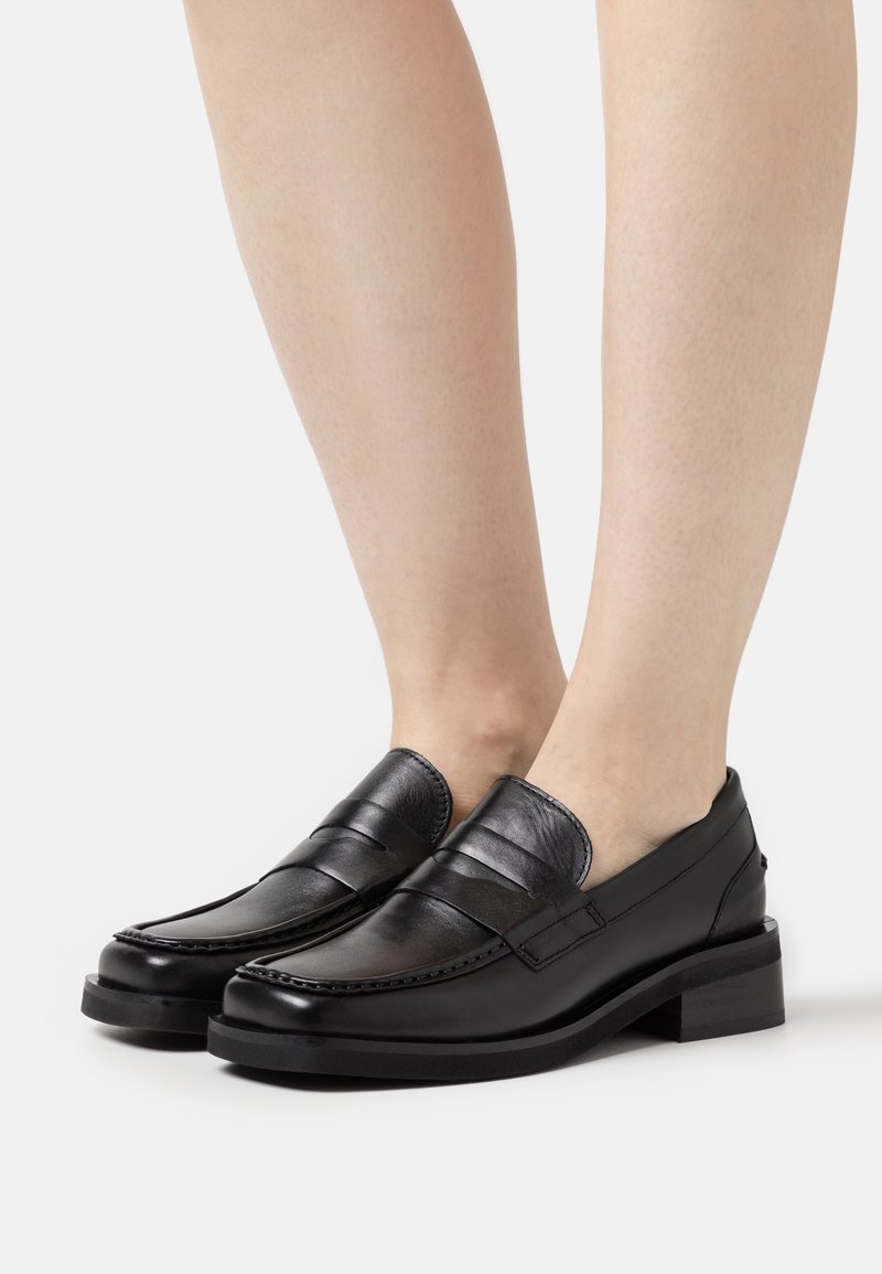 Mocassins en cuir noir avec un petit talon portés pieds nus, présentés de profil sur un fond clair uni.
