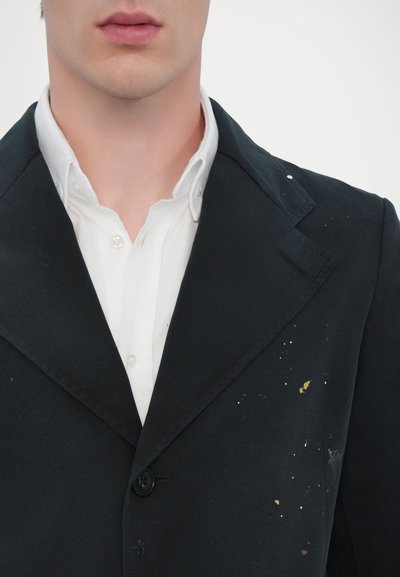 Blazer negro con solapa de muesca, con manchas de pintura amarilla y blanca, llevado sobre una camisa blanca de cuello con botones.