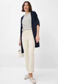 Navy long cardigan over a striped shirt, paired with cream pants featuring a frayed hem, and light loafers with decorative hardware.