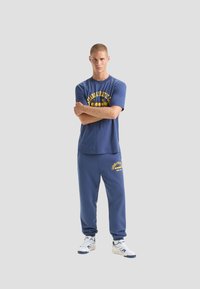 T-shirt di cotone blu con grafica gialla, abbinata a pantaloni jogger blu coordinati con logo. Il modello indossa sneaker bianche. Design semplice e casual.