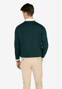 Sudadera de color verde oscuro con cuello redondo y mangas largas, combinada con pantalones beiges. La tela parece suave y tiene un corte clásico.