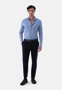 Hellblaues, tailliertes Hemd mit spitzen Kragen, langen Ärmeln, dunkelblauen, maßgeschneiderten Hosen, schwarzem Gürtel und dunklen Schuhen. Glatte Stoffoberfläche.