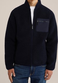 Marinblå fleecejacka med hög krage, full dragkedja och en ficka med dragkedja på bröstet. Mjuk textur med avslappnad passform.