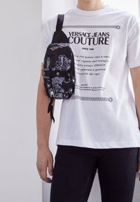 Black fabric crossbody bag with intricate white floral patterns, zippered pocket, adjustable strap, worn over a white printed t-shirt.