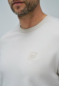 Primer plano de un hombre que lleva un suéter texturizado de color beige claro con un pequeño parche del logo "SALSA JEANS" en el pecho.
