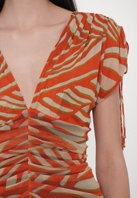 Gros plan d'une femme portant une robe orange et beige à motifs avec un profond décolleté en V et des détails froncés sur l'épaule et la taille.