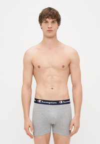 Boxers gris avec une ceinture élastique bleu marine arborant le logo "Champion" en blanc. Tissu doux, design ajusté et longueur mi-cuisse.