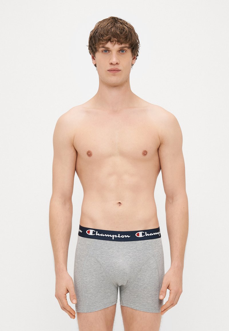 Boxers gris avec une ceinture élastique bleu marine arborant le logo "Champion" en blanc. Tissu doux, design ajusté et longueur mi-cuisse.