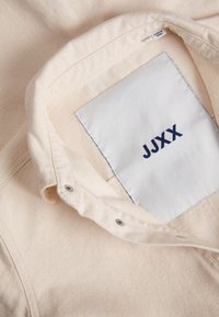 Collar de tela color crema con una etiqueta cosida que muestra "JJXX" en azul oscuro. Presenta costuras visibles y cierres a presión.