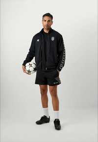 Jachetă sport neagră cu logo și mânecile cu tipar, asortată cu pantaloni scurți negri și ghete. Ținând o minge de fotbal cu accente în formă de stea.