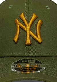 Zelená látková baseballová čiapka s vyšívaným zlatým logom "NY". Obsahuje zaoblenú korunu a okrúhlu nálepku New Era na šiltu.
