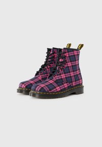 Bottes de combat en tissu écossais noir et rose avec des lacets, surpiqûres jaunes, semelles en caoutchouc et un design de cheville renforcé. Deux languettes de tirage en haut.