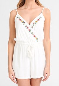 Mono sin mangas blanco con bordado floral a lo largo del escote en V, tirantes finos y cintura con cordón ajustable y borlas.