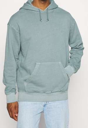 Mand iført lysegrå-grøn hættetrøje med forlomme og snørre, kombineret med lyseblå jeans, stående med den ene hånd i lommen.