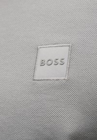 Gros plan sur un tissu texturé gris clair avec une étiquette carrée cousue portant le texte en relief « BOSS » au centre.