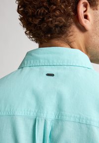 Camicia azzurra chiara realizzata in tessuto testurizzato, con un'etichetta nera sul colletto posteriore e un dettaglio a piega lungo la cucitura.
