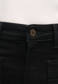 Jeans de ganga pretos com fecho de botão apresentando um logotipo. O tecido tem uma textura suave e inclui bolsos frontais e detalhes cosidos.