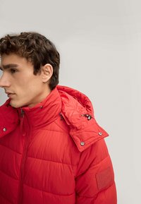 Giacca a vento rossa con colletto alto e cappuccio, caratterizzata da un design trapuntato, chiusura con zip e bottoni a pressione sulle spalle.