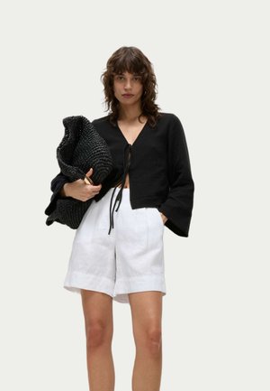 Svart kort blus med utsvängda ärmar och knytning framtill, matchad med vita linnebyxor. Modellen håller en strukturerad svart väska.