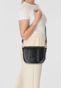 Schwarze Leder-Crossbody-Tasche mit einer gebogenen Klappe, zwei vertikalen Nähten und silbernem Beschlag. Am Schulter getragen mit einem langen Tragegurt.