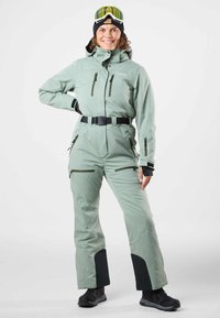 Ski suit in light green, with black accents and adjustable belt. Features zippered pockets and a high collar. Textured fabric, designed for winter sports.