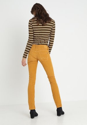 Femme avec des cheveux ondulés portant un pantalon en velours côtelé moutarde, des bottines noires à talons et un haut à manches longues rayé noir, marron et blanc.