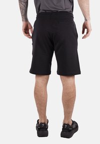 Pantaloni sportivi neri con cintura elastica, texture liscia, design alla lunghezza del ginocchio e dettaglio sulla tasca posteriore. Indossati con sneaker nere.