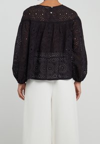 Blouse noire en broderie anglaise avec des manches bouffantes, un col rond et une fermeture à bouton au dos. Présente des découpes détaillées et un tissu texturé.