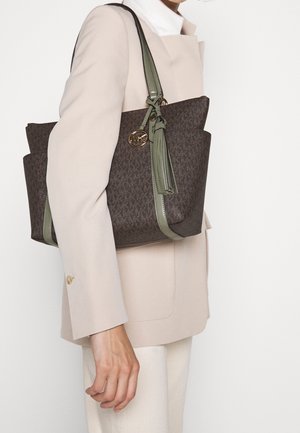 Personne portant un blazer beige et un pantalon clair, portant un grand sac fourre-tout marron à motif avec des anses vertes et un détail de pampille sur l'épaule.