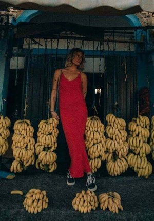 Rotes Spaghettiträgerkleid, getragen von einer Person zwischen hängenden Bündeln gelber Bananen vor einem dunklen Hintergrund mit blauen Akzenten.