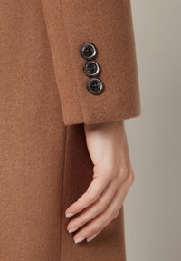 Rękaw brązowego wełnianego płaszcza z trzema czarnymi guzikami przy mankiecie. Miękka tekstura, dopasowany krój i widoczne detale przeszyć.