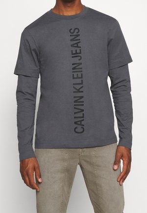 Mężczyzna noszący warstwową ciemnoszarą koszulę z długim i krótkim rękawem z pionowym napisem „CALVIN KLEIN JEANS” oraz jasnoszare spodnie.