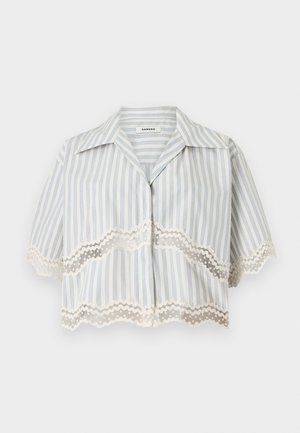 Kvinde står med hånd på hoften iført en hvid stribet bluse med blonde og en navy plisseret nederdel med knapper, brune sandaler.