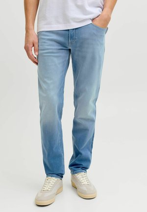 Hombre con vaqueros azul claro, zapatillas blancas y camiseta blanca con una mano en el bolsillo, de pie sobre un fondo claro y sencillo.