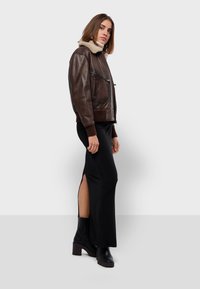 Veste en cuir marron avec un col en shearling beige, dotée de deux poches avant et de poignets côtelés, associée à une robe maxi noire et des bottes noires.