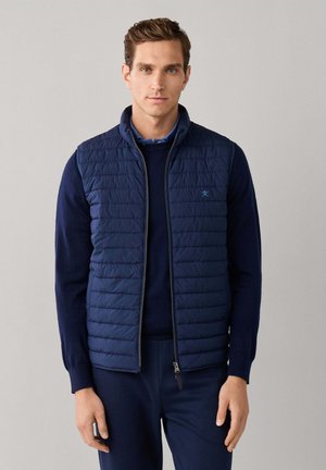 Man standing wearing a navy blue quilted vest over a dark blue sweater and matching pants against a plain light background.
