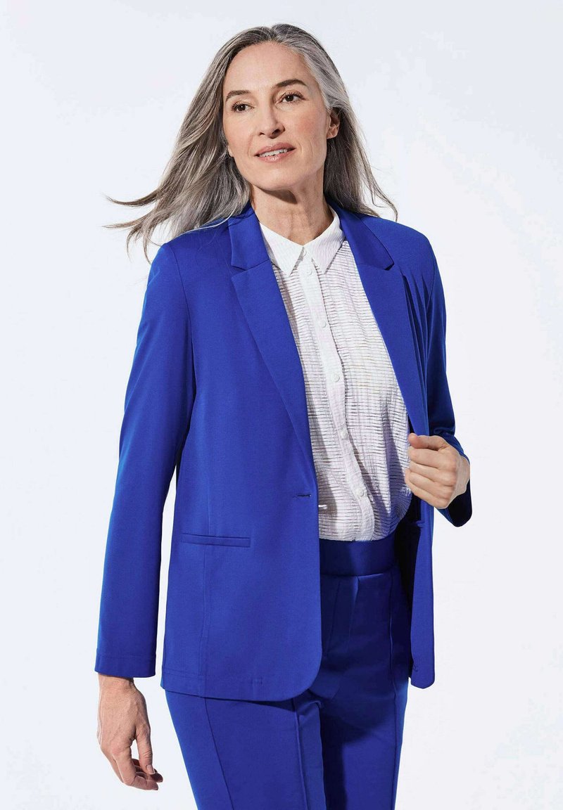 Blazer bleu ajusté avec une texture lisse, doté d'un bouton unique et de deux poches latérales, superposé à une chemise rayée blanche.