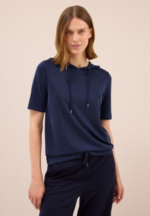 Vrouw met schouderlang lichtbruin haar, gekleed in een kort mouwen marineblauwe hoodie en bijpassende marineblauwe broek tegen een neutrale achtergrond.