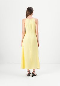 Robe maxi jaune sans manches avec une coupe droite, fermeture éclair au dos et texture lisse. Associée à des sandales à talons noires avec des bretelles.
