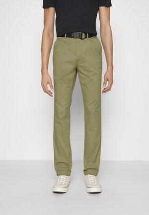 Pantalons en coton chino vert olive avec une coupe droite, dotés de poches latérales et d'une ceinture noire. Portés avec une chemise noire à manches courtes et des baskets.