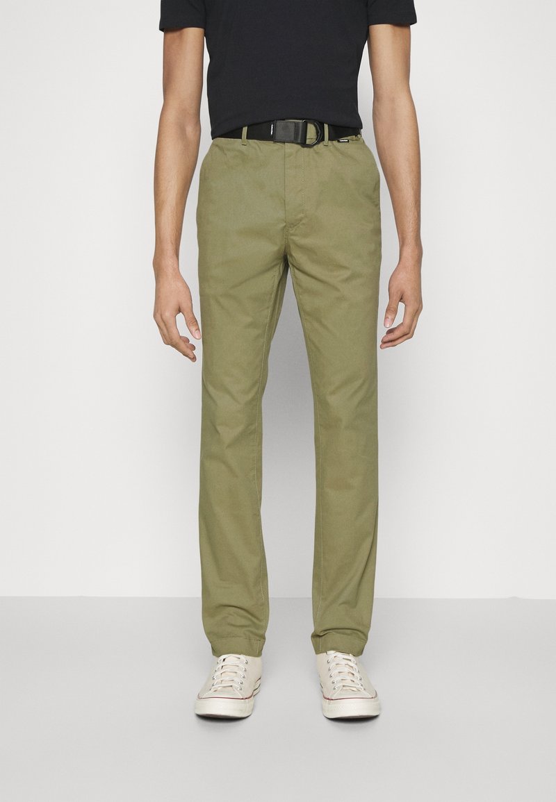 Olijfgroene katoenen chino-broeken met een rechte pasvorm, voorzien van zijzakken en een zwarte riem. Gedragen met een zwart T-shirt met korte mouwen en sneakers.