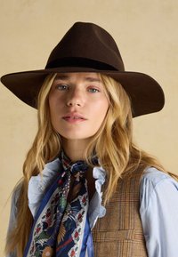 Jeune femme aux longs cheveux blonds portant un chapeau à larges bords brun foncé, un foulard bleu à motifs autour du cou, une chemise à volants et un gilet en tartan marron.
