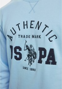 Lyseblå hettegenser med mørk brodert tekst og en polospillergrafikk. Designet inkluderer en ribbestrikket rund hals og dekorativ søm.