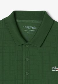 Polo shirt verde con texture e colletto, dotato di una placca con tre bottoni, motivo a griglia sottile e un logo bianco sul lato sinistro del petto.