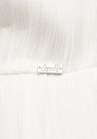 Tela blanca con textura de canalé; una etiqueta rectangular plateada en el centro muestra "Superdry" en estilo cursiva.