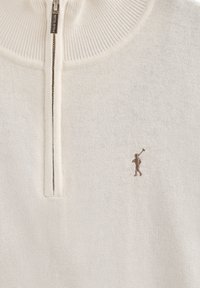 Cremfarbener Strickpullover mit einem Viertel-Reißverschluss am Kragen und einer kleinen, braunen gestickten Figur auf der linken Brust. Weicher, strukturierten Stoff.