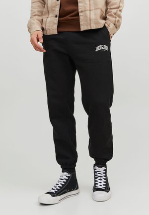 Mann trägt schwarze Jack & Jones Jogginghose, schwarze High-Top-Sneaker mit weißen Schnürsenkeln und eine beige karierte Jacke mit den Händen in den Taschen.