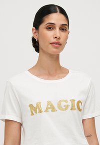 White cotton t-shirt featuring the word "MAGIC" in bold gold glitter font. Classic crew neckline and rolled short sleeves. Smooth texture.