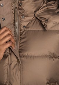 Giacca a vento marrone con finitura lucida, dotata di zip frontale e bottoni a pressione, che presenta sezioni trapuntate e un'etichetta in tessuto.