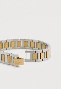 Nerūsējošā tērauda aproce ar zelta toņa akcentiem, kurai ir fakturēti taisnstūrveida posmi un aizdares mezgls ar reljefā izceltu logotipu.